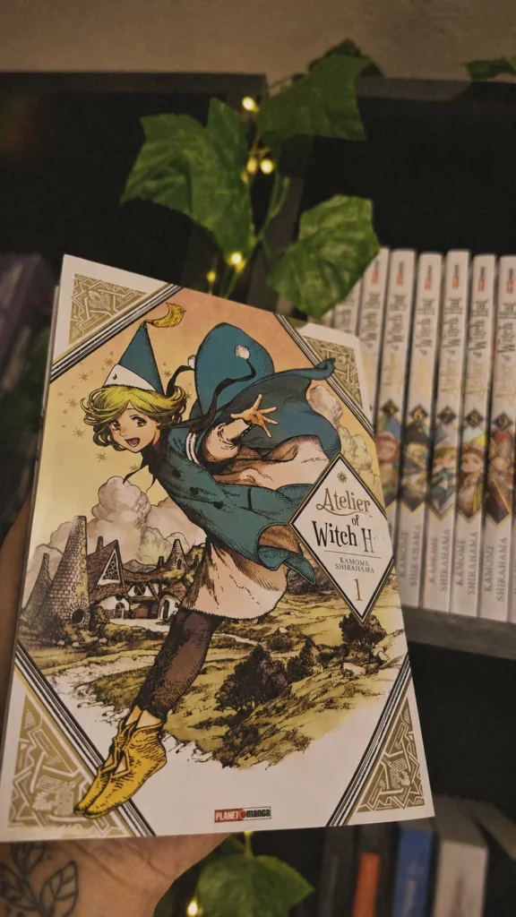 o volume 1 de witch hat atelier, de kamome shirahama, segurado na mão diante da estante